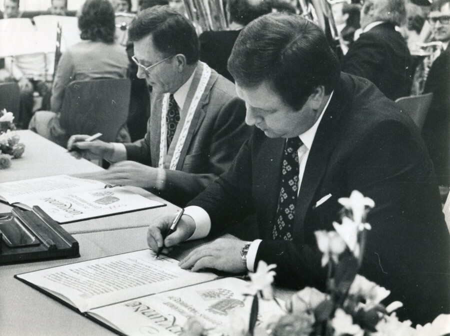 Das Foto zeigt Pierre Quéméner (†, hinten) mit Holzwickedes Bürgermeister Heinrich Schürhoff (vorn) bei der Unterzeichnung der Partnerschaftsurkunde. (Foto: (Foto: Gemeindearchiv)